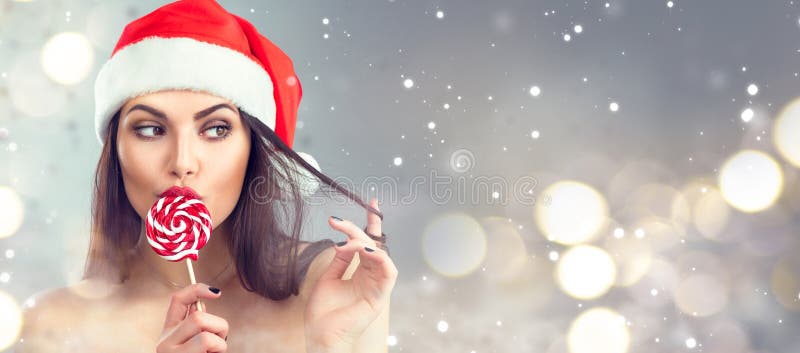 Christmas woman. Beauty model girl in Santa Claus hat with red lips and xmas lollipop candy. Closeup portrait