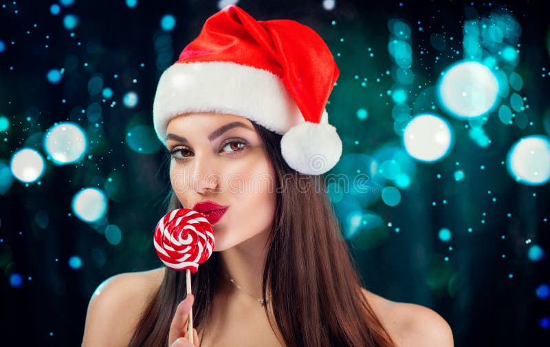 Christmas woman. Beauty model girl in Santa Claus hat with red lips and xmas lollipop candy in her hand. Joy. Surprised expression