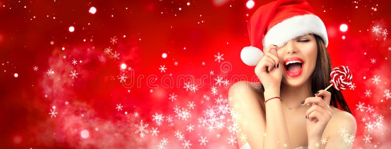 Christmas woman. Joyful model girl in Santa`s hat with red lips and lollipop candy in her hand