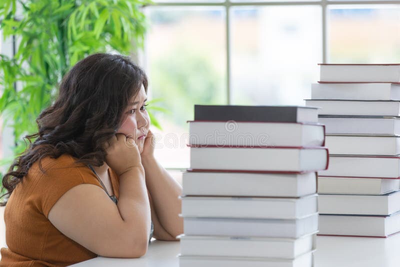 Chubby woman student sitting and looking on a stack of books on a table with anxious in a room of the house. Study and work from