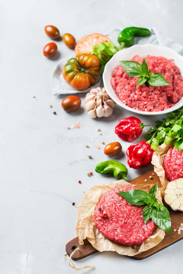 Chuleta y verduras picaditas orgánicas crudas hechas en casa de la hamburguesa de la carne de la carne de vaca. Comida sana, cocinando concepto Carne de la carne de vaca y chuleta picaditas orgánicas crudas hechas en casa del filete de la hamburguesa con las verduras en una tabla blanca