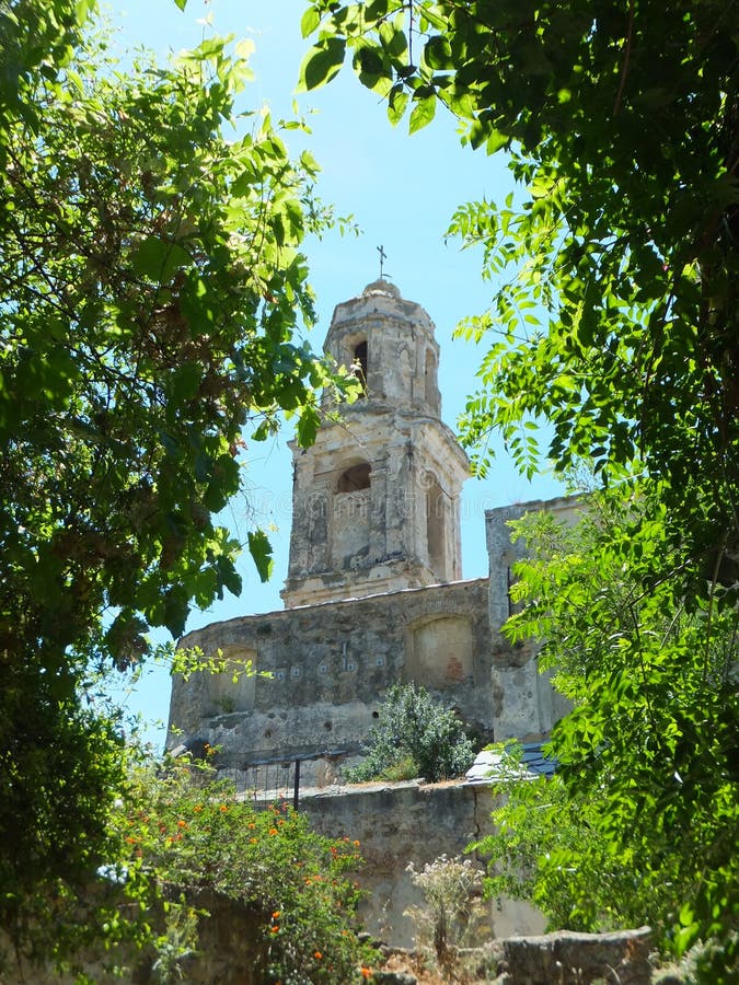 Church spire in Bussana Vecchia seen in an opening between green leafy tree branches on a sunny day