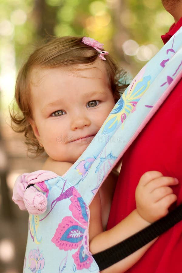 Happy baby sitting in a carrying sling. Father carry a child comfortable. Walking outdoors. Selective focus on the face of the baby. Happy baby sitting in a carrying sling. Father carry a child comfortable. Walking outdoors. Selective focus on the face of the baby.