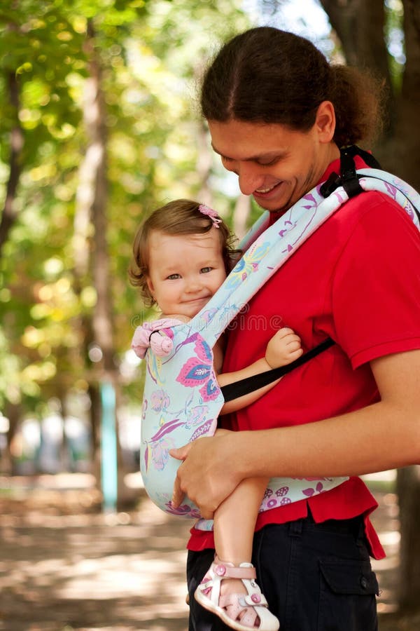 Happy baby sitting in a carrying sling. Funny father and baby walking outdoors. Father carry a child comfortable. Selective focus on the face of the baby. Happy baby sitting in a carrying sling. Funny father and baby walking outdoors. Father carry a child comfortable. Selective focus on the face of the baby.