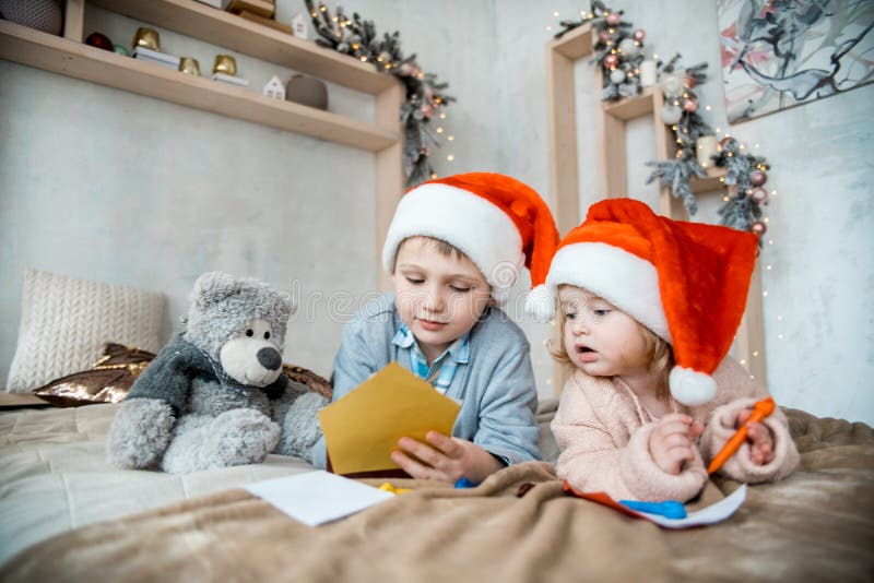 Chłopiec i dziewczynka w Santa hat pisze list do Świętego Mikołaja Przytulny, jasny salon Drzewo bożonarodzeniowe i światła fotografia stock Chłopiec i dziewczynka w Santa hat pisze list do Świętego Mikołaja Przytulny, jasny salon Drzewo bożonarodzeniowe i światła fotografia stock