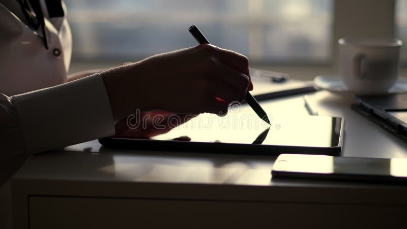 dark silhouette of businesswoman, closeup of hands. she writes something in tablet, next is a laptop on table. Blurred window background , a ray of light is reflected in the tablet screen. High quality photo. dark silhouette of businesswoman, closeup of hands. she writes something in tablet, next is a laptop on table. Blurred window background , a ray of light is reflected in the tablet screen. High quality photo