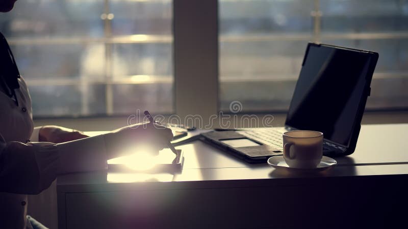 dark silhouette of businesswoman, closeup of hands. she writes something in tablet, next is a laptop on table. Blurred window background , a ray of light is reflected in the tablet screen. High quality photo. dark silhouette of businesswoman, closeup of hands. she writes something in tablet, next is a laptop on table. Blurred window background , a ray of light is reflected in the tablet screen. High quality photo