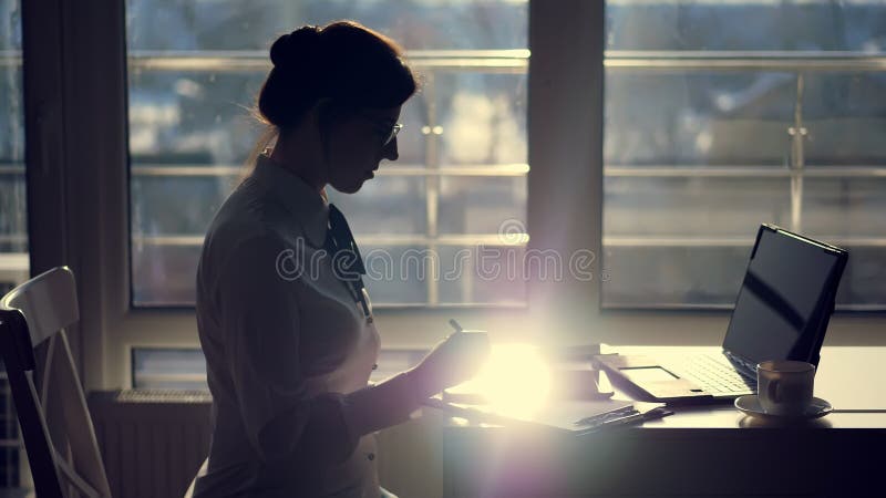 dark silhouette of Young Focused attentive businesswoman wearing glasses, writes something in tablet, next is a laptop on table. against the window, a ray of light is reflected in the tablet screen. High quality photo. dark silhouette of Young Focused attentive businesswoman wearing glasses, writes something in tablet, next is a laptop on table. against the window, a ray of light is reflected in the tablet screen. High quality photo