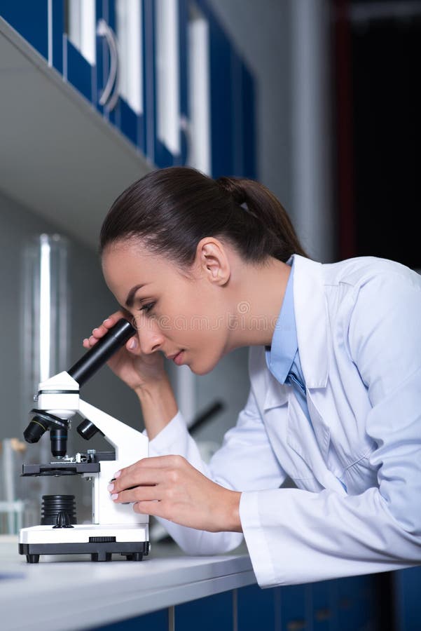 Científico bonito joven que se coloca y que mira en el microscopio fotos de archivo libres de regalías Científico bonito joven que se coloca y que mira en el microscopio fotos de archivo libres de regalías