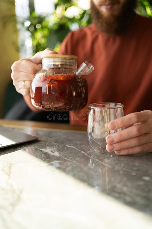 Cierre de las manos macho vertiendo té de fruta de una tetera de vidrio en una taza sobre una mesa de madera fotografía de archivo