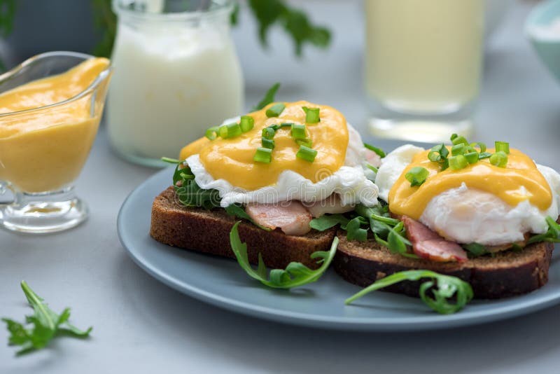 Cierre encima de los huevos Benedicto con arugula, salsa del tocino y del hollandaise en fondo de madera gris Foco suave Concepto imagen de archivo libre de regalías Cierre encima de los huevos Benedicto con arugula, salsa del tocino y del hollandaise en fondo de madera gris Foco suave Concepto imagen de archivo libre de regalías