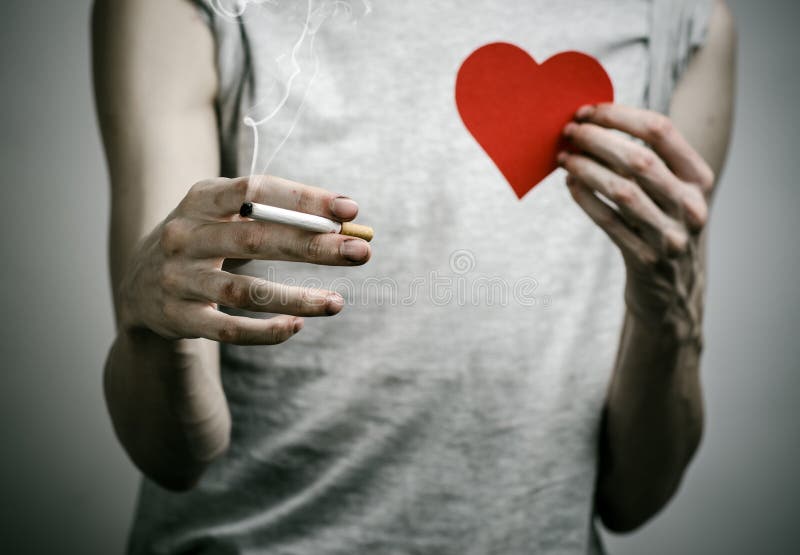Cigarettes, addiction and public health topic: smoker holds the cigarette in his hand and a red heart on a dark background in the. Cigarettes, addiction and stock photo Cigarettes, addiction and public health topic: smoker holds the cigarette in his hand and a red heart on a dark background in the. Cigarettes, addiction and stock photo