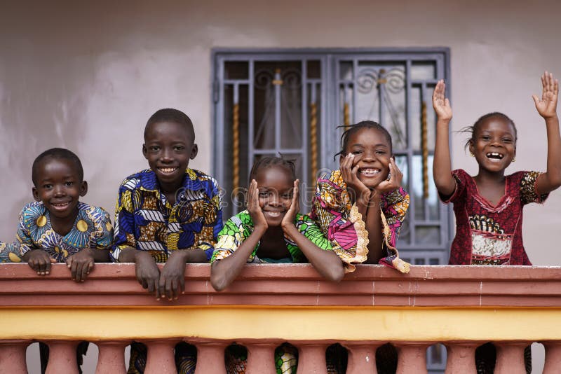 Cinco crianças africanas saudando transeuntes de uma varanda caseira colonial