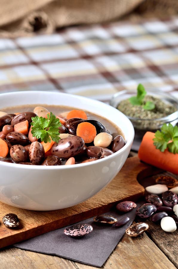 Bowl of bean soup with large beans on cutting board, carrots, parsley, marjoram, spoon and ladle, towel in background - vertical photo. Bowl of bean soup with large beans on cutting board, carrots, parsley, marjoram, spoon and ladle, towel in background - vertical photo