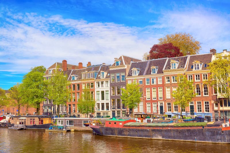 Cityscape mening van het kanaal van Amsterdam in de zomer met een blauwe hemel, huisboten en traditionele oude huizen Schilderach stock afbeelding Cityscape mening van het kanaal van Amsterdam in de zomer met een blauwe hemel, huisboten en traditionele oude huizen Schilderach stock afbeelding