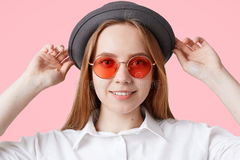 Ciérrese encima del retrato de la hembra encantada preciosa con sonrisa agradable, lleva las gafas de sol rojas elegantes y el so foto de archivo Ciérrese encima del retrato de la hembra encantada preciosa con sonrisa agradable, lleva las gafas de sol rojas elegantes y el so foto de archivo