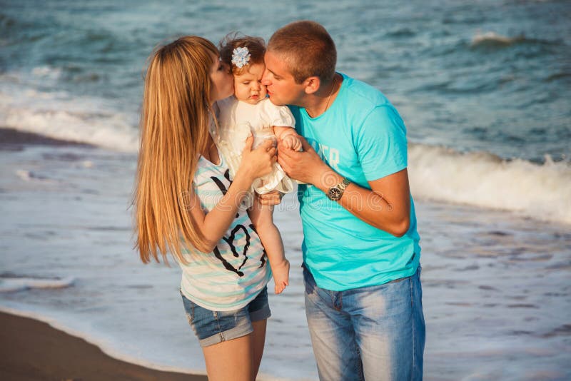 Ciérrese para arriba de la familia cariñosa feliz joven que abraza y que besa a la pequeña hija en la playa junto cerca del océan fotografía de archivo Ciérrese para arriba de la familia cariñosa feliz joven que abraza y que besa a la pequeña hija en la playa junto cerca del océan fotografía de archivo