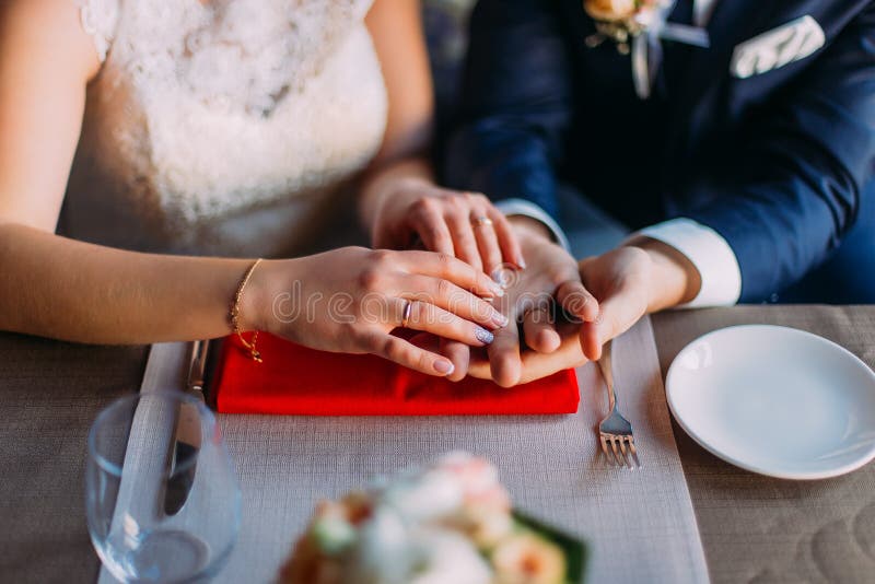 Ciérrese para arriba de las manos nuevo-casadas de un par que llevan los anillos de bodas en la tabla servida con artículos de co fotografía de archivo libre de regalías Ciérrese para arriba de las manos nuevo-casadas de un par que llevan los anillos de bodas en la tabla servida con artículos de co fotografía de archivo libre de regalías