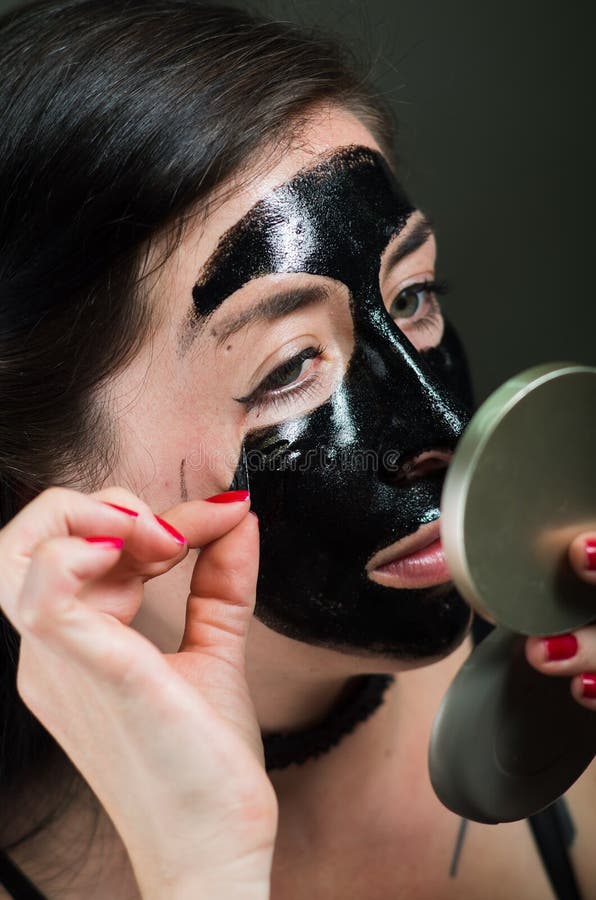 Ciérrese para arriba de una mitad del lanzamiento de la mujer joven de la belleza de una mascarilla negra que mira el espejo foto de archivo libre de regalías Ciérrese para arriba de una mitad del lanzamiento de la mujer joven de la belleza de una mascarilla negra que mira el espejo foto de archivo libre de regalías