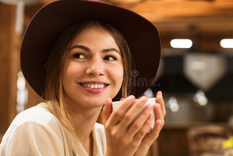 Ciérrese para arriba de una muchacha feliz en el sombrero que se sienta en la tabla del café dentro imagen de archivo libre de regalías Ciérrese para arriba de una muchacha feliz en el sombrero que se sienta en la tabla del café dentro imagen de archivo libre de regalías