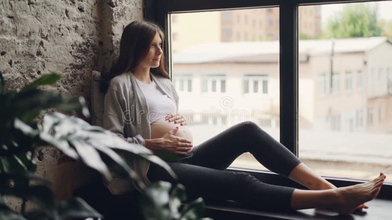 Ciężarnej dziewczyny piękna brunetka jest siedząca na nadokiennym parapecie, uderzanie jej brzuch i patrzeć z okno odpoczywać zdjęcie wideo Ciężarnej dziewczyny piękna brunetka jest siedząca na nadokiennym parapecie, uderzanie jej brzuch i patrzeć z okno odpoczywać zdjęcie wideo