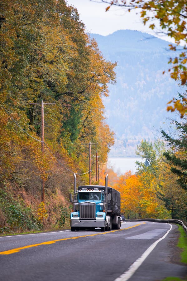 Large classic blue semi truck with tarp covered trailer climbing uphill on a scenic highway with metal security fence and road markings surrounded by yellow autumn trees on both sides of the road on the background of the river and mountains. Large classic blue semi truck with tarp covered trailer climbing uphill on a scenic highway with metal security fence and road markings surrounded by yellow autumn trees on both sides of the road on the background of the river and mountains
