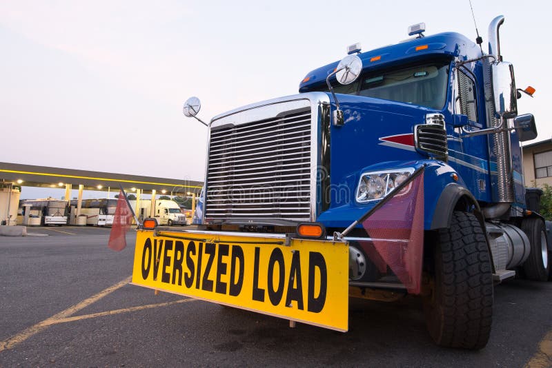 Classic blue truck with the sign oversized load on the truck stop on the background of a gas station with truck and buses. Front view opens up a powerful vehicle with a grille, the turned wheel, the side signal flags and rear-view mirrors, attached to the hood. Classic blue truck with the sign oversized load on the truck stop on the background of a gas station with truck and buses. Front view opens up a powerful vehicle with a grille, the turned wheel, the side signal flags and rear-view mirrors, attached to the hood.