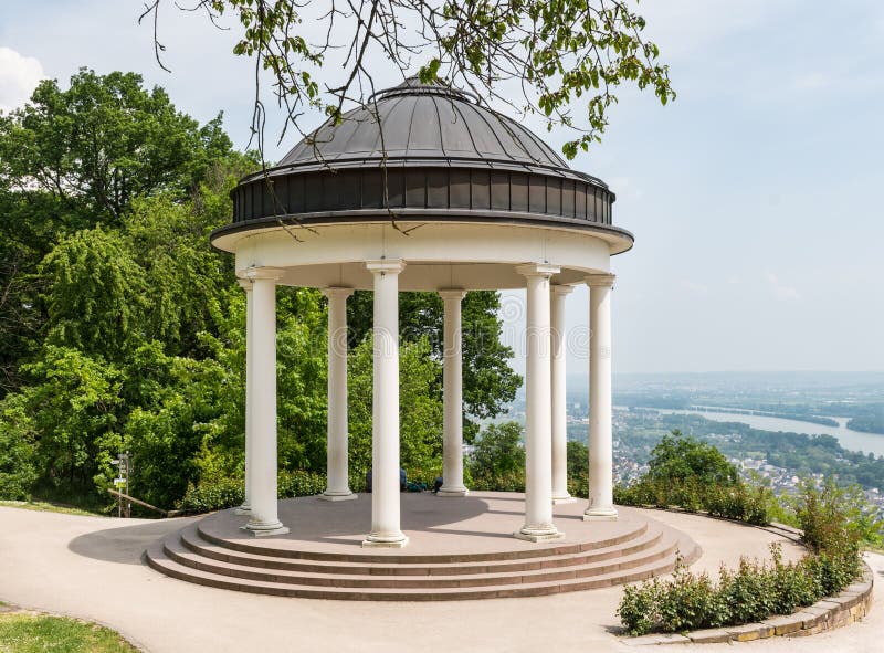 This is a serene view of a classical white pavilion situated atop a hill above Rudesheim in Germany, offering a panoramic vista of the river Rhine below under a clear summer sky. This is a serene view of a classical white pavilion situated atop a hill above Rudesheim in Germany, offering a panoramic vista of the river Rhine below under a clear summer sky