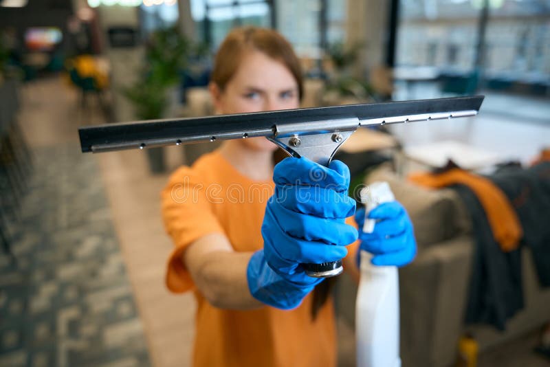 Cleaning lady in uniform and protective gloves uses glass scraper and spray to work. Cleaning lady in uniform and protective gloves uses glass scraper and spray to work