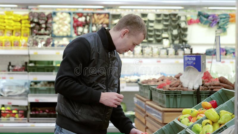 Cliente con una chaqueta negra inspecciona un pimiento en una estantería de un supermercado evaluando cuidadosamente la vibrante s almacen de video