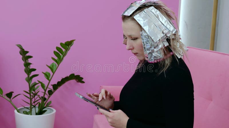 Cliente esperando en la peluquería mientras se ponen tintes usando un smartphone. la escena captura un momento de multitarea en un almacen de video