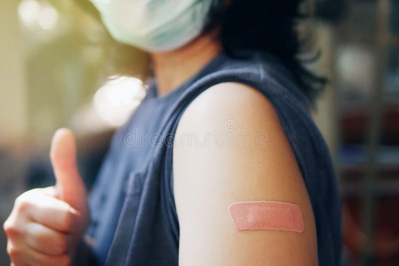 Close-up adhesive bandage on unrecognized person`s arm after injection of vaccine, people in face mask received a coronavirus. COVID-19 vaccine and giving thumb