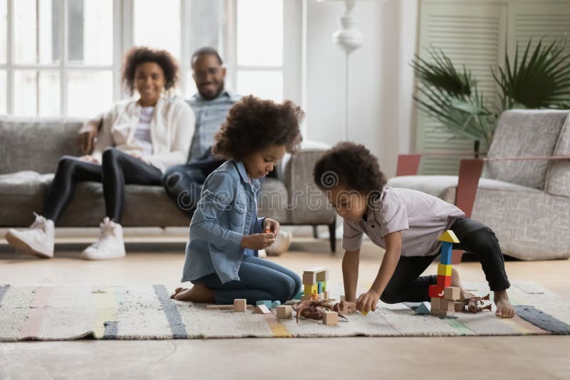 Close up African American kids, sister and brother playing together while parents relaxing on couch, cute little girl and boy sitting on warm floor, building tower, using colorful wooden blocks. Close up African American kids, sister and brother playing together while parents relaxing on couch, cute little girl and boy sitting on warm floor, building tower, using colorful wooden blocks