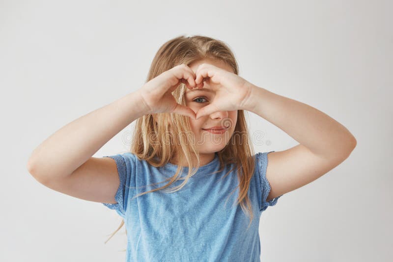 Close up of beautiful blonde girl making heart with hands, looking through it in camera, posing for photo with smile and