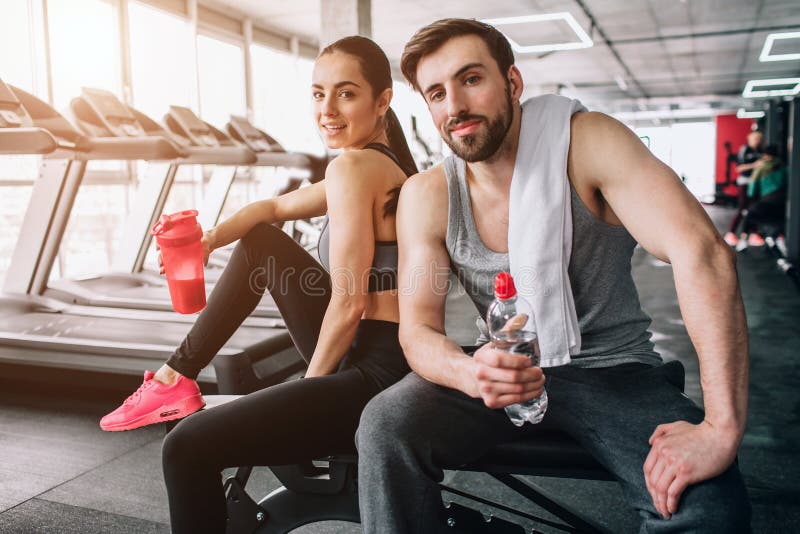 Close up of a beautiful couple sitting on the sport bench and posing. Also they are drinking water from their bottles and having rest after hard workout