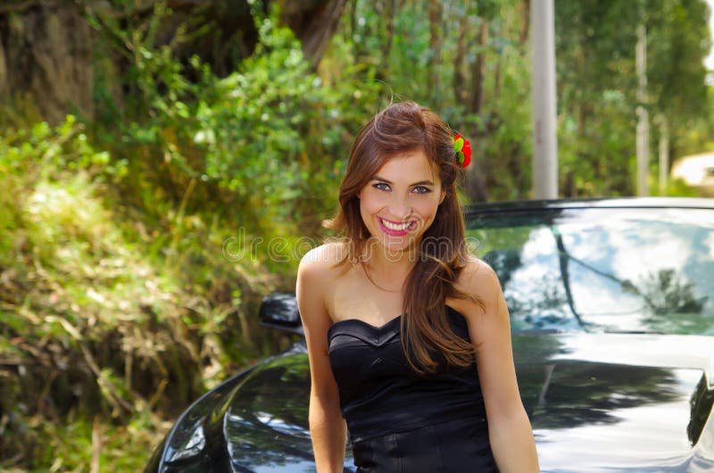 Close up of beautiful woman wearing a black dress and a red flower in her head and posing in front of a luxury black car