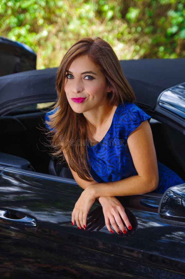 Close up of beautiful woman wearing a blue dress and posing inside of a luxury black car on a roadtrip. the car standing