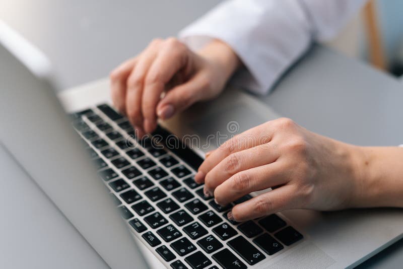 Close-up cropped shot of unrecognizable thoughtful female doctor wearing white medical uniform using laptop keyboard sitting at desk in modern office of medic clinic, cropped shot, high angle view.