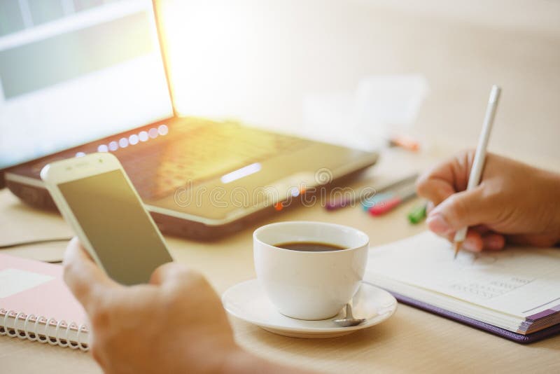 Close up cup of coffee and laptop computer with hand of business man using smart phone.