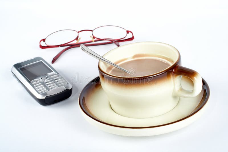 Close-up of a cup of coffee with the spoon inside, cellular phone and pair of glasses on white background