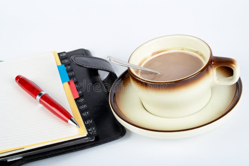 Close-up of a cup of coffee with the spoon inside and notebook with ballpoint on white background