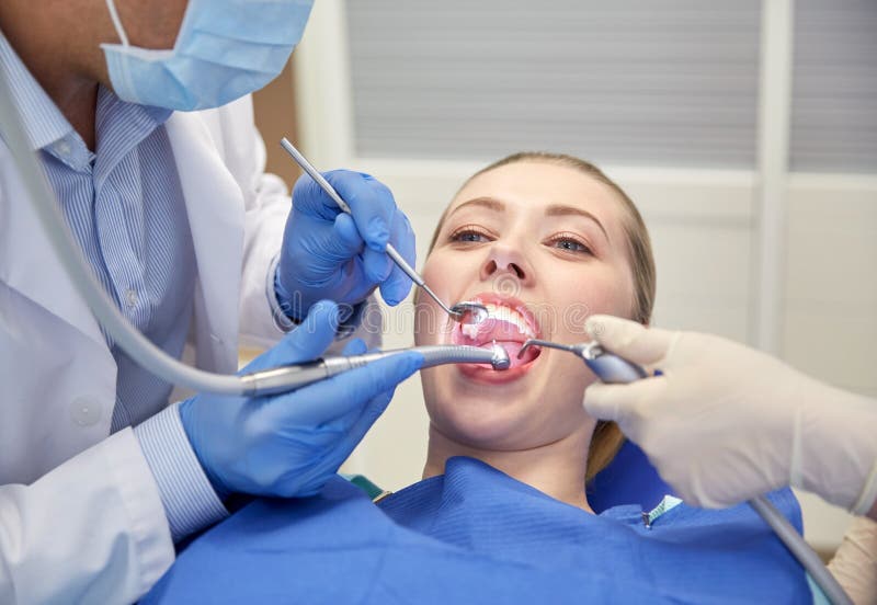 People, medicine, stomatology and health care concept - close up of dentists and assistant with mirror, drill and dental air water gun spray treating female patient teeth at dental clinic. People, medicine, stomatology and health care concept - close up of dentists and assistant with mirror, drill and dental air water gun spray treating female patient teeth at dental clinic