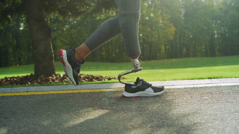 Close up disabled woman legs in sports shoes running on road. Girl feet training outdoors in slow motion. Female jogger legs doing cardio workout in park. Runner legs running outside. Close up disabled woman legs in sports shoes running on road. Girl feet training outdoors in slow motion. Female jogger legs doing cardio workout in park. Runner legs running outside