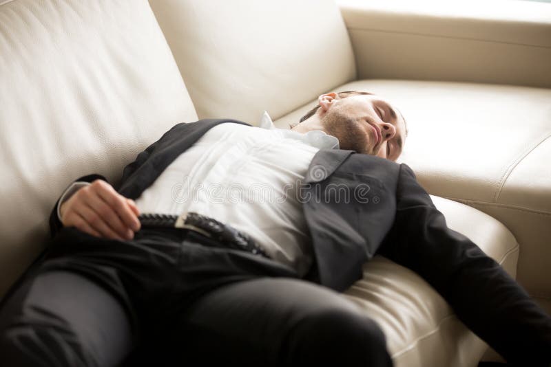 Close up of exhausted businessman laying on the couch. Young hard working entrepreneur fell asleep on sofa at workplace. Staying at work overtime, overworked, too much workload, lack of sleep concept.