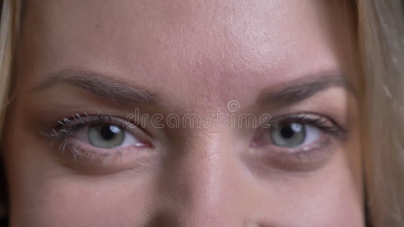 Close-up eyes portrait of middle-aged blonde businesswoman watches contentedly into camera on black background.