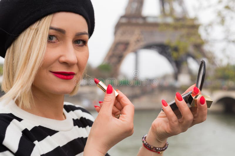 Close-up of face of fashionable blonde who paints her lips with red lipstick, against background of Eiffel Tower in Paris on spring day. Walking around city. Beauty and fashion. Tourism and travel. Close-up of face of fashionable blonde who paints her lips with red lipstick, against background of Eiffel Tower in Paris on spring day. Walking around city. Beauty and fashion. Tourism and travel.