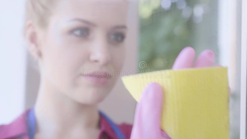 Close-up face of pretty smiling blond girl washing the window with window rag in the room. Cleaning day. Housekeeping