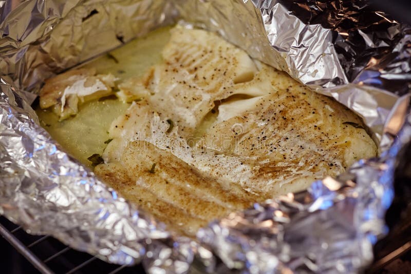 Close up fillet of white fish in the foil, sprinkled with black pepper and aromatic herbs, on a baking sheet removed from the oven