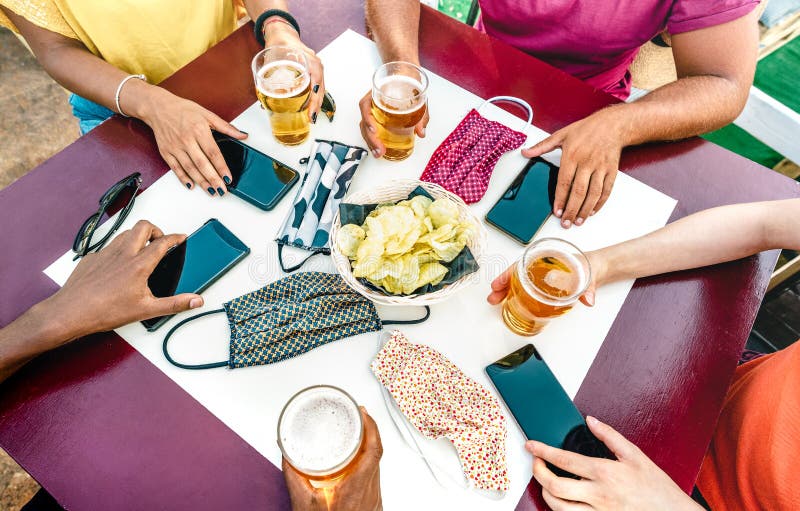 Close up of friends hands near face masks on table with mobile smart phones and beers - New normal lifestyle technology concept. With people at party bar royalty free stock images