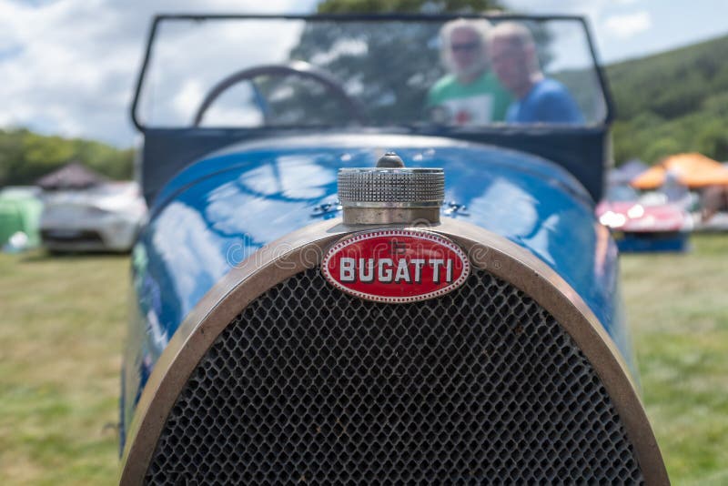 A close up of the front of an old Bugatti car with its red emblem with two older gentlemen admiring the car in the